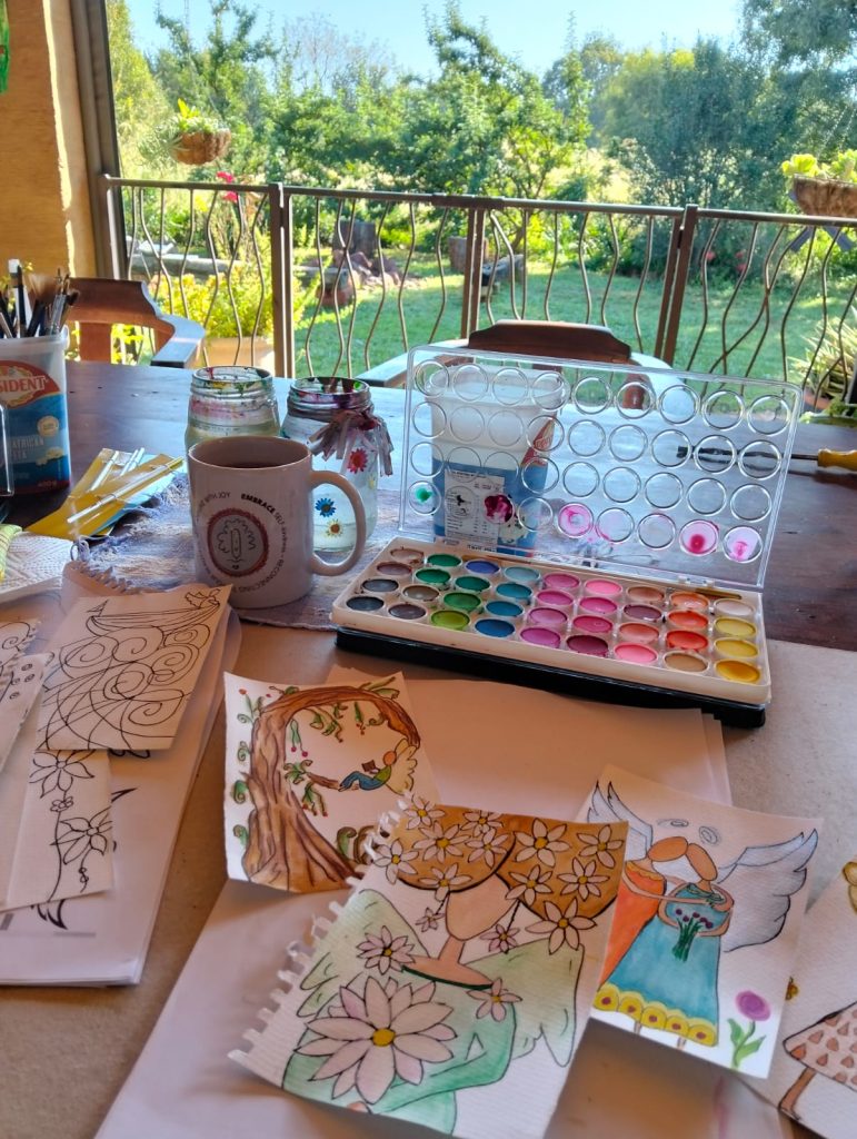 Hand-painted angel card in progress on the stoep at Lekker Plekkie, with paintbrushes and coffee nearby. hand-painted-angel-card-set-nellvanderloo-lekker-plekkie-buy-online